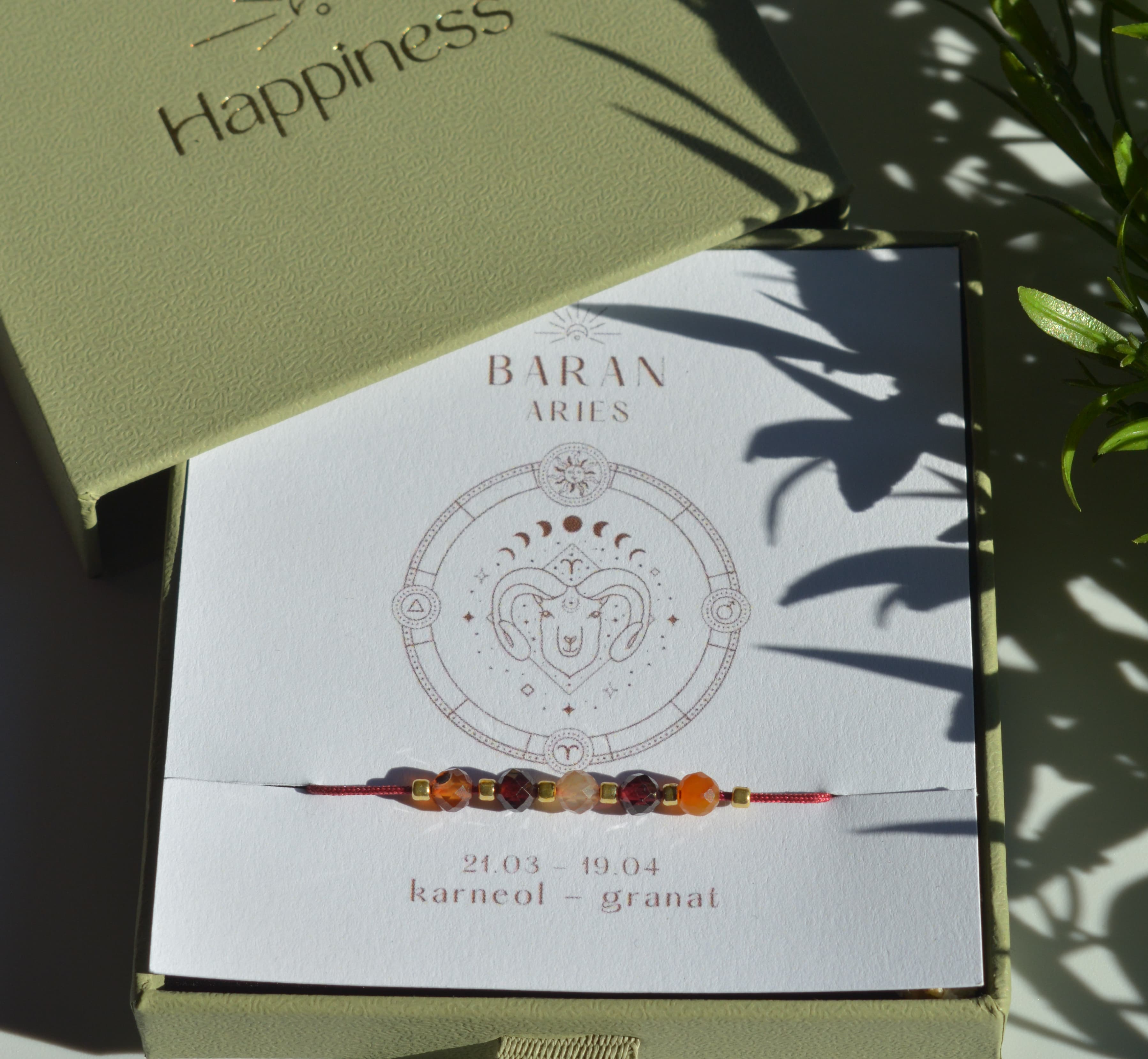 Bransoletka na czerwonym sznurku nylonowym z koralikami z kamieni naturalnych: pomarańczowym karneolem i ciemnoczerwonym granatem. Opakowana w pudełko w kolorze oliwkowym ze złotym logo Happiness.
