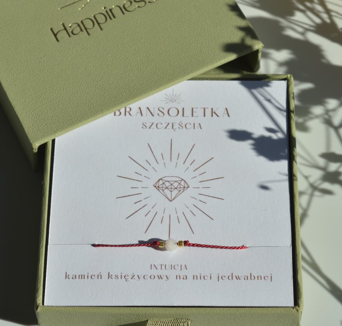 Bransoletka szczęścia na ciemnoczerwonej nici jedwabnej. W centralnej części koralik kamienia księżycowego i dwa malutkie szklane koraliki w kolorze złotym. Opakowana w oliwkowe pudełko ze złotym logo happiness.