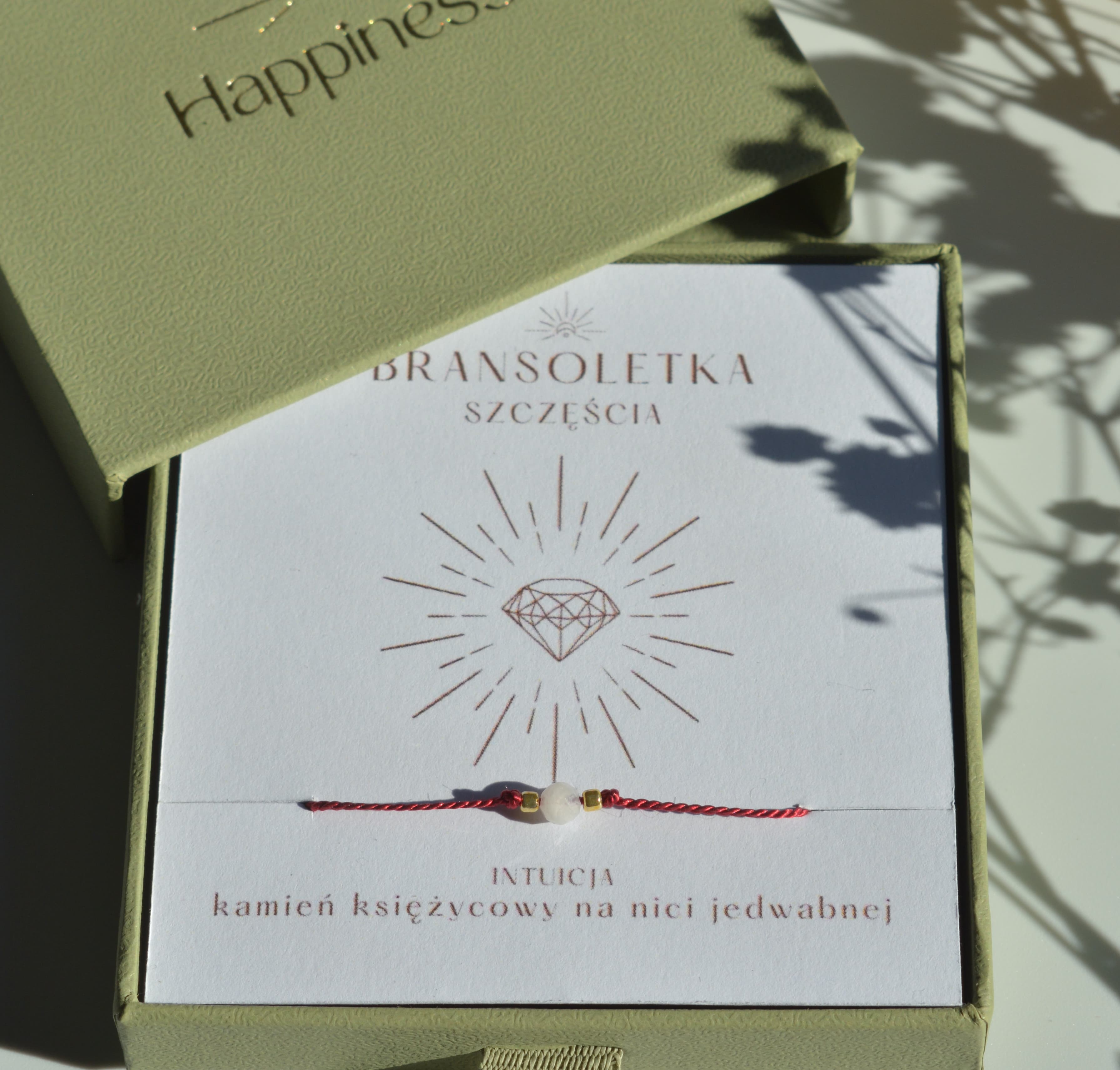 Bransoletka szczęścia na ciemnoczerwonej nici jedwabnej. W centralnej części koralik kamienia księżycowego i dwa malutkie szklane koraliki w kolorze złotym. Opakowana w oliwkowe pudełko ze złotym logo happiness.