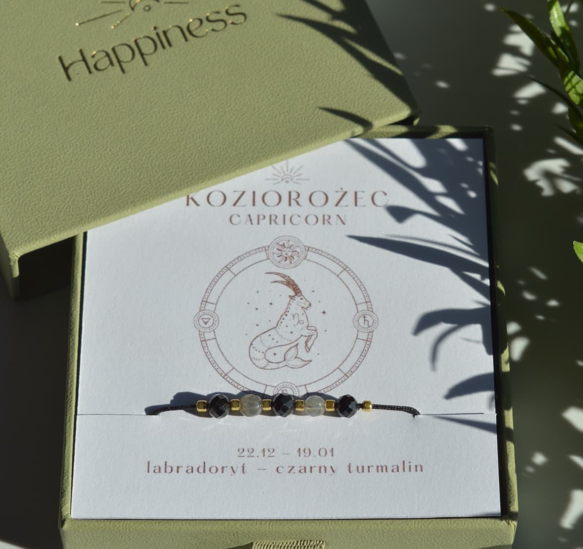 Bransoletka na czarnym sznurku nylonowym z koralikami z kamieni naturalnych: szarym labradorytem i czarnym turmalinem. Opakowana w pudełko w kolorze oliwkowym ze złotym logo Happiness.
