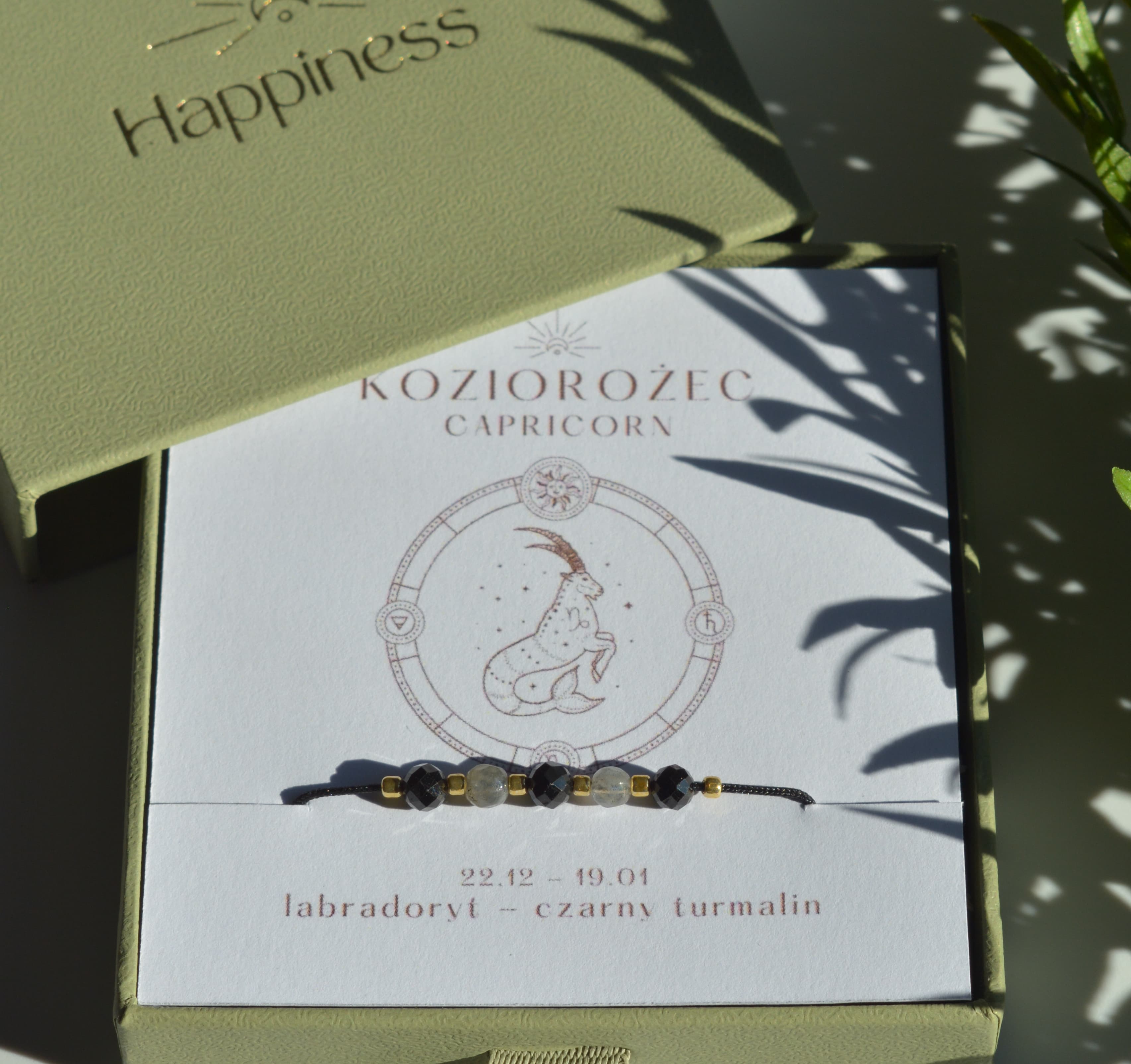 Bransoletka na czarnym sznurku nylonowym z koralikami z kamieni naturalnych: szarym labradorytem i czarnym turmalinem. Opakowana w pudełko w kolorze oliwkowym ze złotym logo Happiness.
