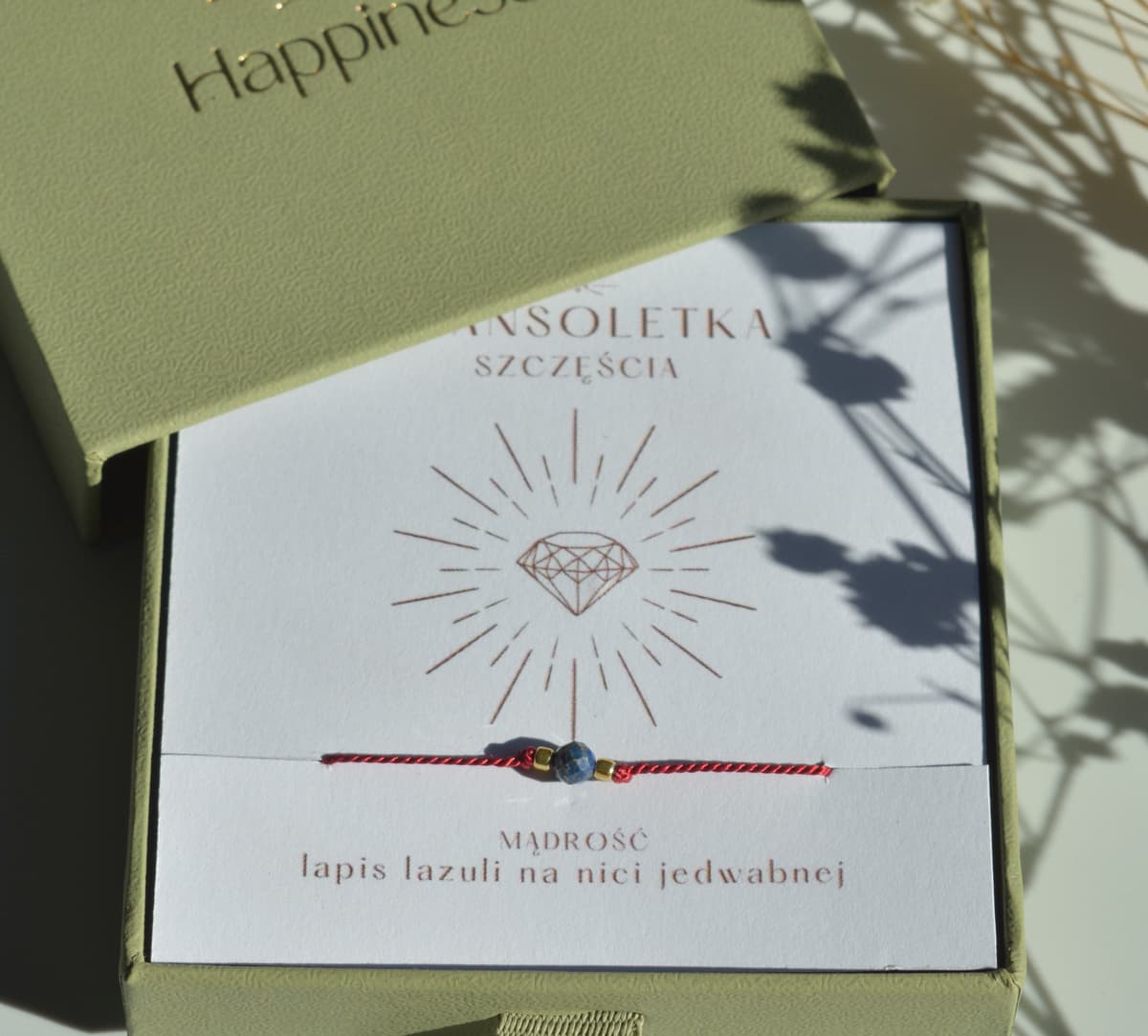 Bransoletka szczęścia na ciemnoczerwonej nici jedwabnej. W centralnej części koralik lapis lazuli i dwa malutkie szklane koraliki w kolorze złotym. Opakowana w oliwkowe pudełko ze złotym logo happiness.