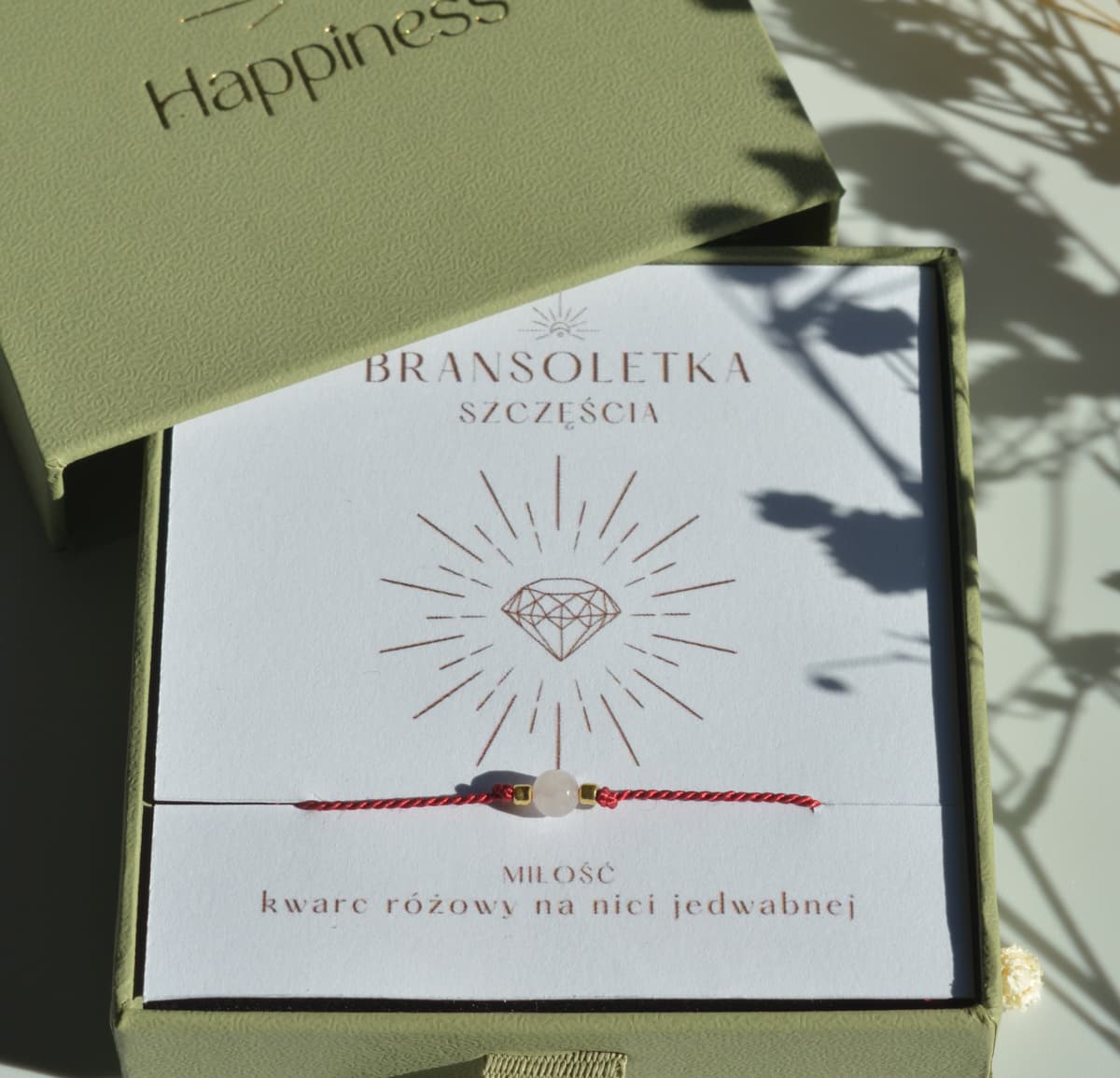 Bransoletka szczęścia na ciemnoczerwonej nici jedwabnej. W centralnej części koralik różowego kwarcu i dwa malutkie szklane koraliki w kolorze złotym. Opakowana w oliwkowe pudełko ze złotym logo happiness.