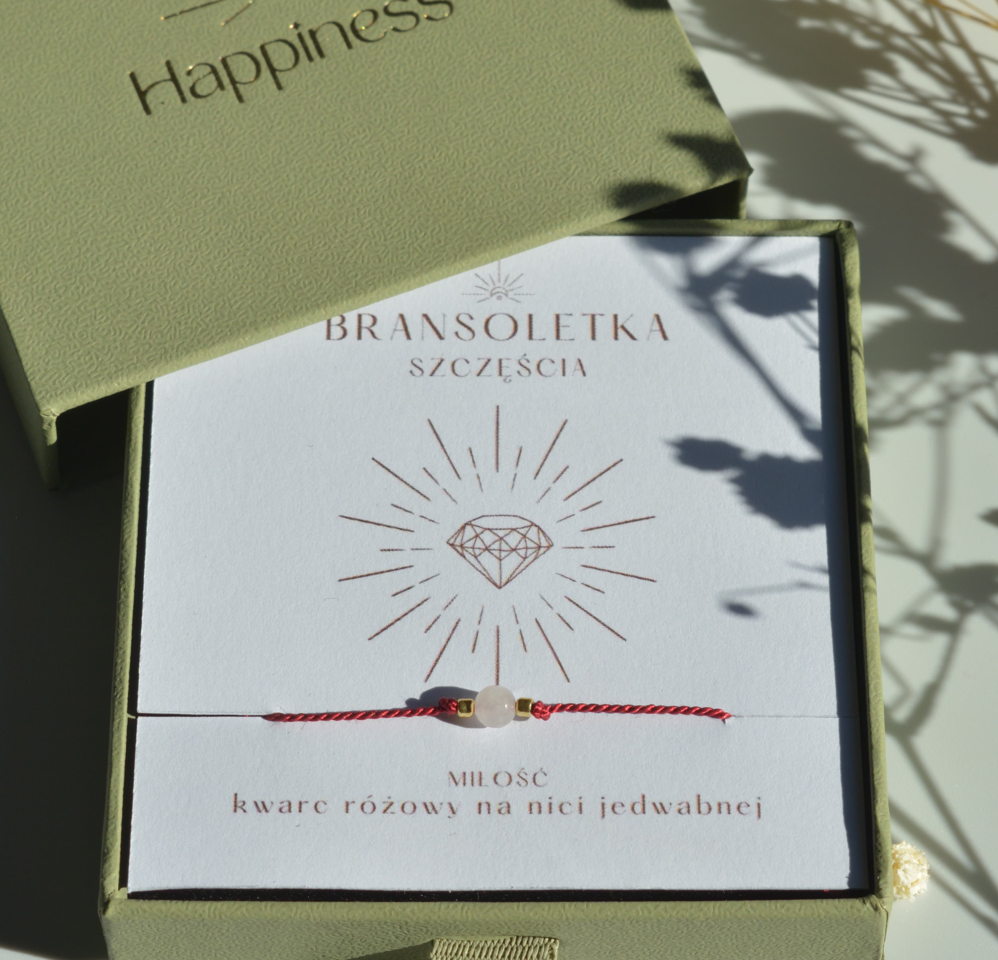 Bransoletka szczęścia na ciemnoczerwonej nici jedwabnej. W centralnej części koralik różowego kwarcu i dwa malutkie szklane koraliki w kolorze złotym. Opakowana w oliwkowe pudełko ze złotym logo happiness.