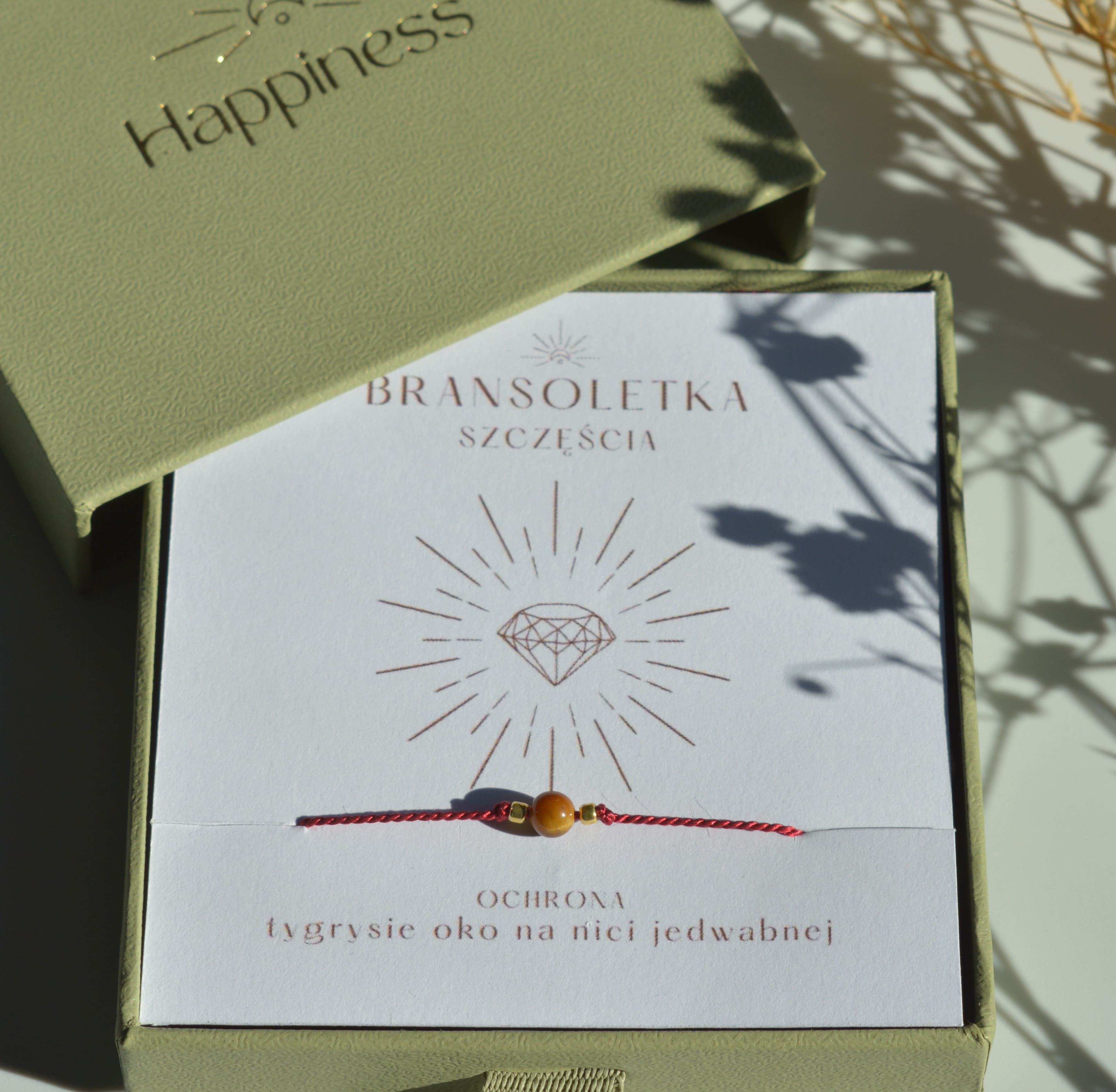 Bransoletka szczęścia na ciemnoczerwonej nici jedwabnej. W centralnej części koralik tygrysiego oka i dwa malutkie szklane koraliki w kolorze złotym. Opakowana w oliwkowe pudełko ze złotym logo happiness.