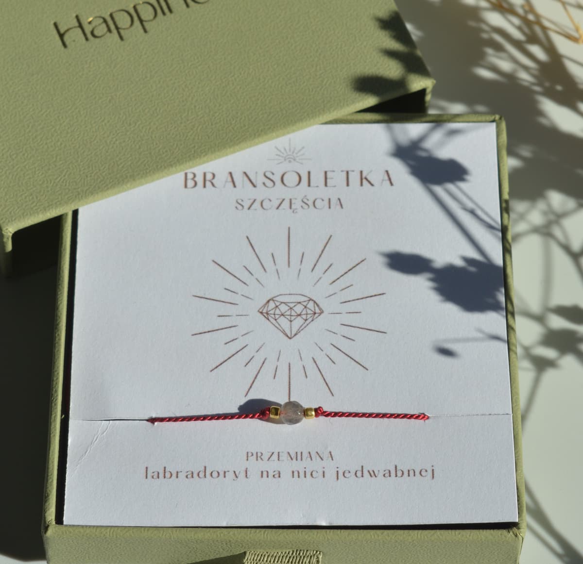 Bransoletka szczęścia na ciemnoczerwonej nici jedwabnej. W centralnej części koralik labradorytu i dwa malutkie szklane koraliki w kolorze złotym. Opakowana w oliwkowe pudełko ze złotym logo happiness.