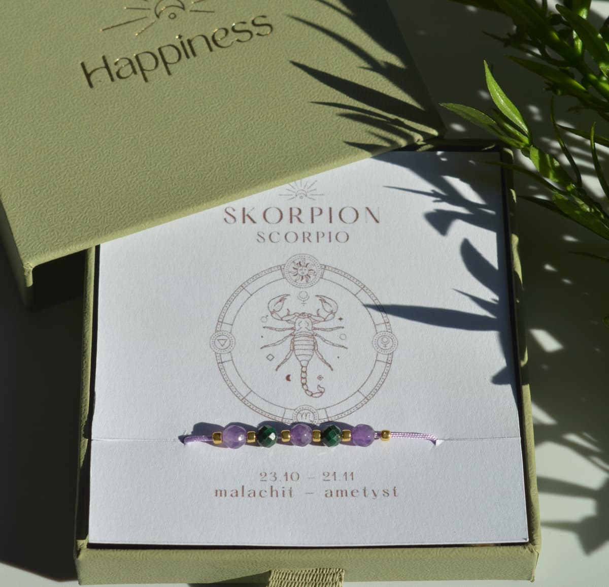 Bransoletka na fioletowym sznurku nylonowym z koralikami z kamieni naturalnych: ciemnozielonym malachitem i fioletowym ametystem. Opakowana w pudełko w kolorze oliwkowym ze złotym logo Happiness.