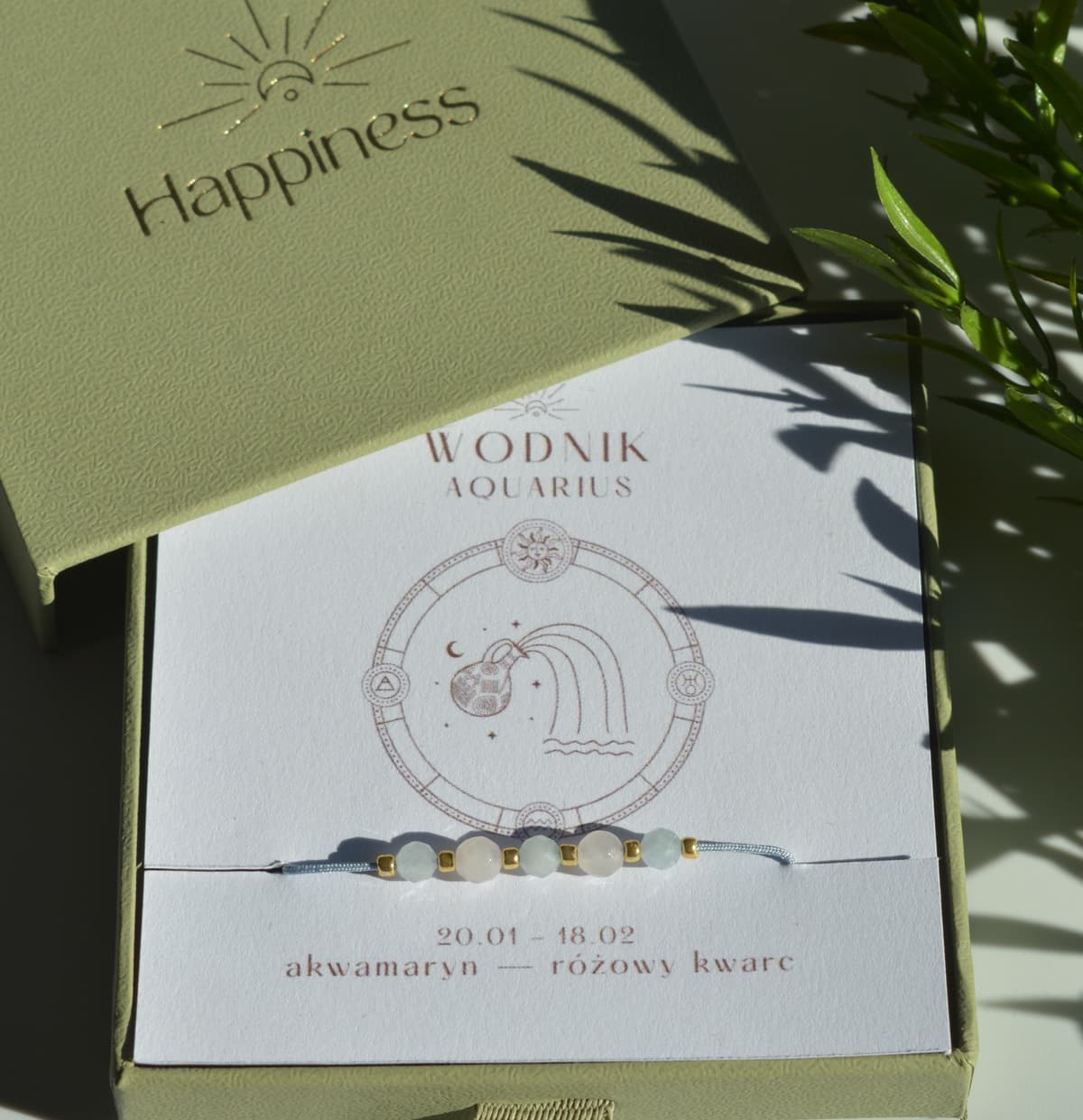 Bransoletka na błękitnym sznurku nylonowym z koralikami z kamieni naturalnych: błękitnym akwamarynem i różowym kwarcem. Opakowana w pudełko w kolorze oliwkowym ze złotym logo Happiness.